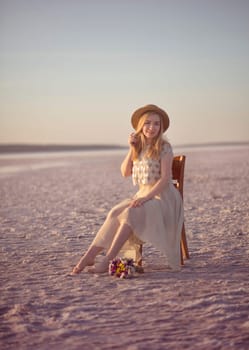 Blonde girl with a straw hat sits in the middle of a salt lake. Her dress flutters in the wind. The setting sun illuminates the shore covered with salt. Kuyalnik, Odessa