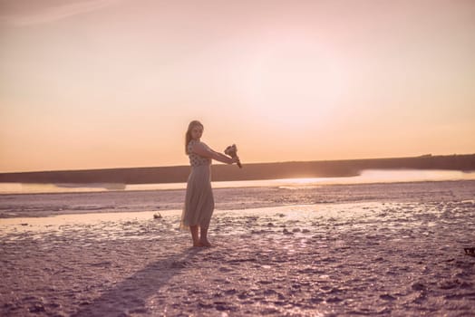 blonde girl stands in the middle of a salt lake. her dress flutters in the wind. a bright circle of the setting sun illuminates the shore covered with salt. she holds a bouquet in her hands. kuyalnik, odessa