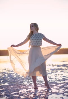 Slender blonde girl stands on the surface of sea salt at sunset. She is illuminated by the setting sun. Her chiffon dress is illuminated. Her silhouette on the horizon. Kuyalnik, Odessa, Ukraine