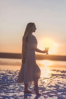 Slender blonde girl stands on the surface of sea salt at sunset. She is illuminated by the setting sun. Her chiffon dress is illuminated. She has a glass in her hand. Salt lake on Kuyalnik in Odessa
