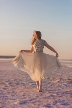 Slender blonde girl stands on the surface of sea salt at sunset around the salt lake. General plan, she is illuminated by the setting sun, her chiffon dress flutters. in the photo there is a salt lake