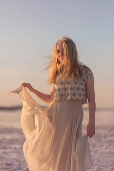 Slender blonde girl stands on the surface of sea salt at sunset. She is illuminated by the setting sun. Her chiffon dress flutters. She is standing on the salt lake on Kuyalnik in Odessa
