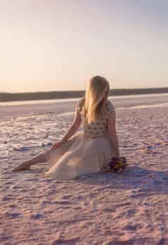 blonde girl sits in the middle of a salt lake. her dress flutters in the wind. the setting sun illuminates the shore covered with salt. kuyalnik, odessa