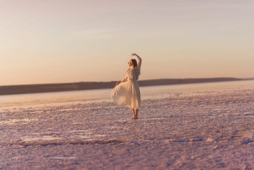 Slender blonde girl stands on the surface of sea salt at sunset around the salt lake. General plan, she is illuminated by the setting sun, her chiffon dress flutters. She is dancing. In the photo there is a salt lake, Kuyalnik, Odessa