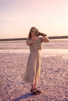 blonde girl stands in the middle of a salt lake. her dress flutters in the wind. the surface is pink. she has a bouquet in her hands, she covers her face. kuyalnik, odessa