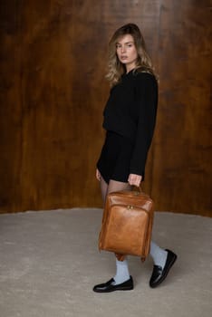 A girl in a warm sweater, a black short skirt, nylon tights, black loafers and gray socks is posing with a brown backpack