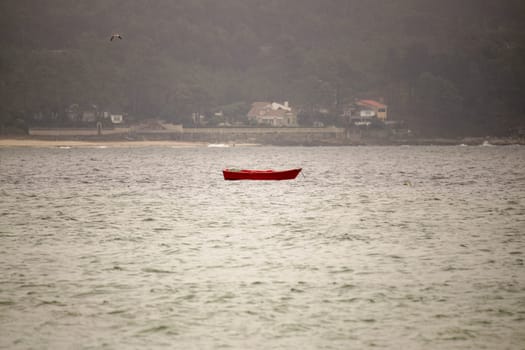 A vivid red boat gently floats on the tranquil ocean, surrounded by fog, creating a serene atmosphere reflecting peace and solitude amid visible coastal homes.