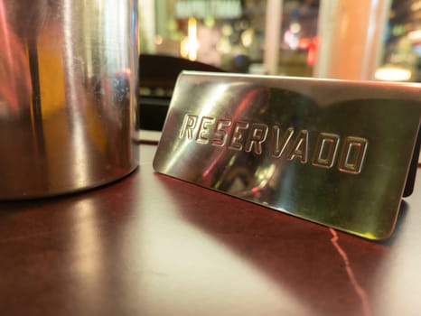 A close-up view of a metal reservado sign placed on a table, suggesting a reserved seating area in a vibrant restaurant or cafe atmosphere with blurred lights.