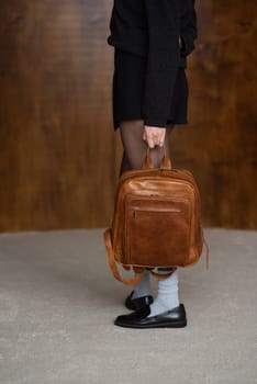 A girl in a warm sweater, a black short skirt, nylon tights, black loafers and gray socks is posing with a brown backpack