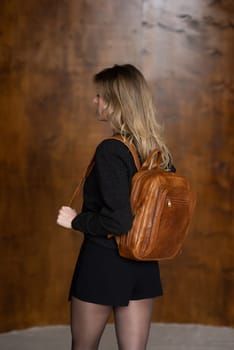 A girl in a warm sweater, a black short skirt and nylon tights is posing with a light brown backpack