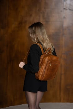 A girl in a warm sweater, a black short skirt and nylon tights is posing with a light brown backpack