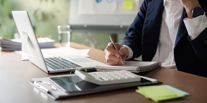 An elderly businessman writes notes while reviewing financial documents and using a calculator in a business setting.