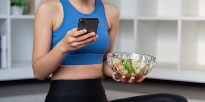 A fit woman in activewear checks her smartphone while holding a bowl of fresh salad, embodying a healthy lifestyle.