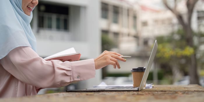 A Muslim woman in a hijab engages with her laptop and takes notes in an outdoor workspace, highlighting her productivity and organization.
