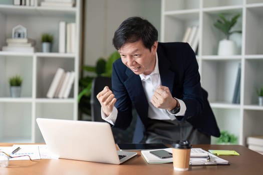 An elderly businessman expresses excitement and triumph after achieving a milestone while working on his laptop.