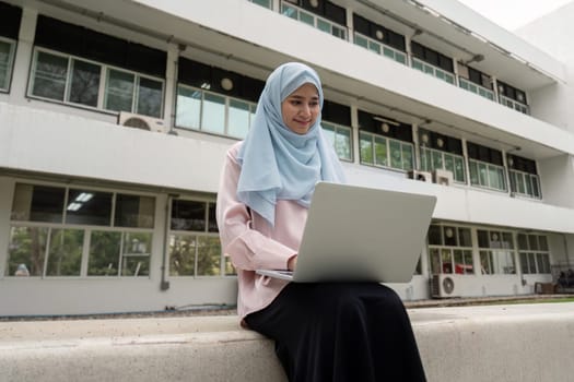 A Muslim woman in a hijab sits outdoors, focused on her laptop while engaging in business activities, showcasing professionalism.