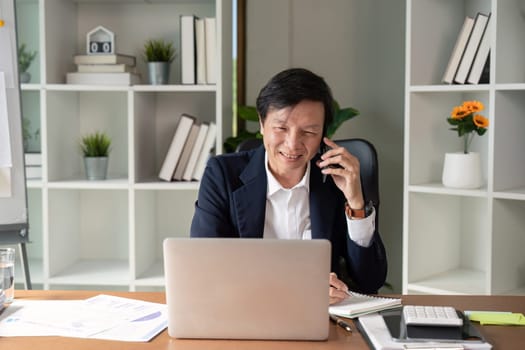An elderly businessman engages in a phone conversation while reviewing financial documents in a contemporary office setting.