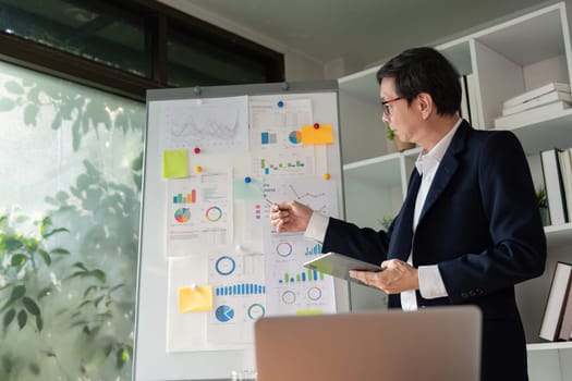 An elderly businessman stands by a board presenting financial data, highlighting analytical skills in a contemporary workspace.