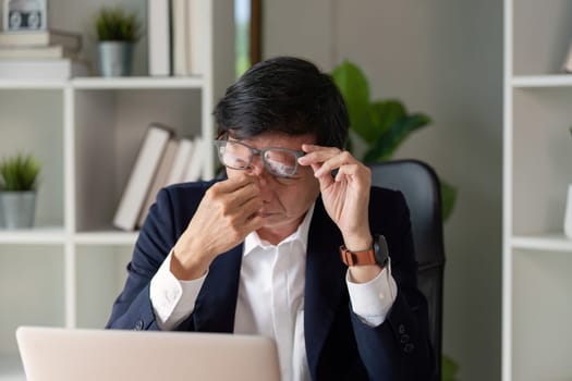 An elderly businessman rubs his eyes in frustration while working on a laptop, showcasing the challenges of modern work life.