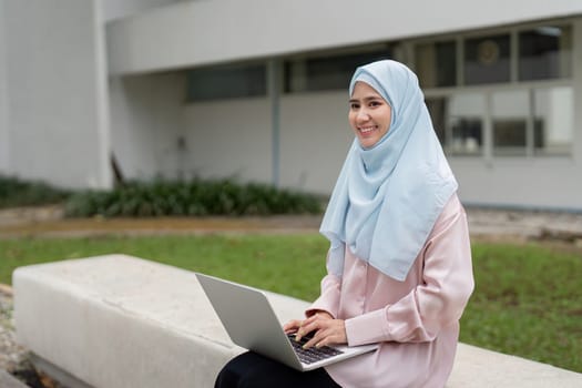 A joyful Muslim woman in a hijab is working on her laptop outside, promoting a positive and productive work environment.
