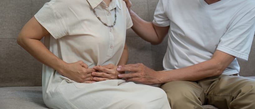 An elderly couple on a couch, one partner holding their abdomen in discomfort while the other offers support and care.