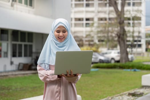 A joyful Muslim woman in a hijab smiles while using her laptop outdoors, showcasing positivity in her business endeavors.