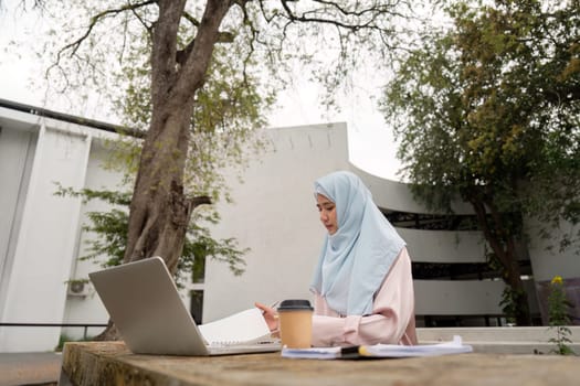 A Muslim woman in a hijab studies outdoors with her laptop and notes, illustrating her commitment to learning and professional growth.