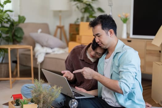 A gay couple sits together, working on a laptop while unpacking boxes in their new home, showcasing teamwork and love.