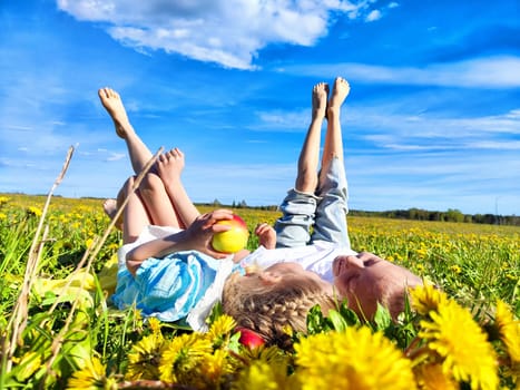 Two children lie on their backs in a vibrant dandelion field, enjoying fresh air and a bright sunny day while holding an apple.