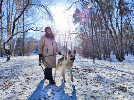 A woman walks her German Shepherd dog along a snowy path, enjoying the fresh air and winter sunshine in a peaceful park setting.
