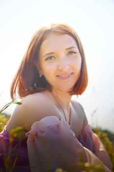 A young woman enjoys a sunny afternoon, smiling while sitting in a vibrant green field, with her hair gently illuminated by sunlight.