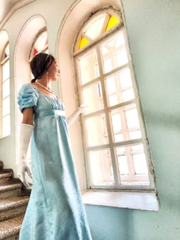A girl in a vintage blue dress gazes out of a brightly lit window in a historic castle, embodying a romantic princess's spirit.