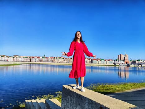A woman smiles joyfully while standing on a concrete ledge near a river, showcasing vibrant architecture and clear blue sky.