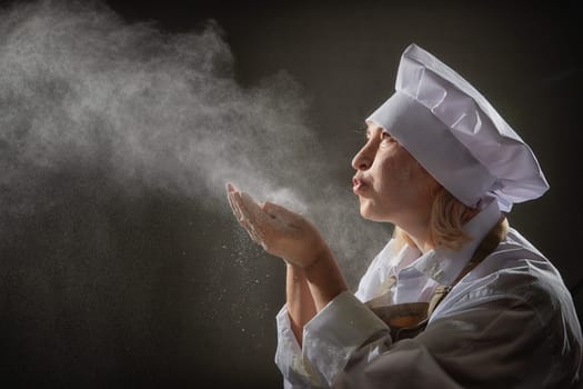 An elderly woman dressed as a cook joyfully blows flour while enjoying a lighthearted kitchen photoshoot with whimsical expressions.