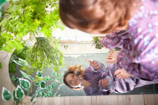 The woman is engaged in a moment of reflection, sitting thoughtfully near a mirror surrounded by vibrant plants.
