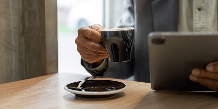 An elderly man enjoys a cup of coffee while reviewing business documents on a tablet, illustrating a modern approach to work life balance.