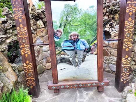 A mature woman takes a humorous selfie while posing in a curved mirror surrounded by nature in an outdoor park setting.