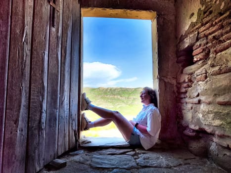 A middle-aged woman takes a break in a picturesque doorway, enjoying her travel journey while capturing moments for her blog.