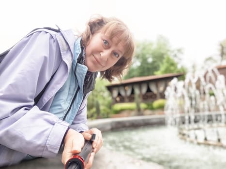 In a serene park setting, a middle-aged woman captures moments while taking selfies near a peaceful fountain.