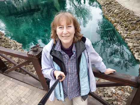 A middle-aged woman with blonde hair captures selfies in a beautiful park during her trip, surrounded by lush nature.