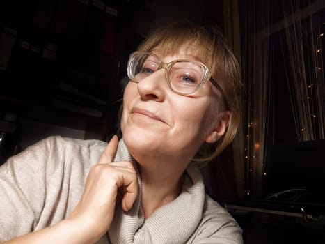 A middle-aged woman with blonde hair is posing for a selfie while looking relaxed and happy in her cozy home setting.