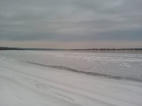 Amidst the calm of winter, the riverbank transforms into a serene landscape where snow covers the ground, evoking a sense of quiet reflection. The soft hues of the cloudy sky meet the horizon, creating a stunning juxtaposition of water and white.