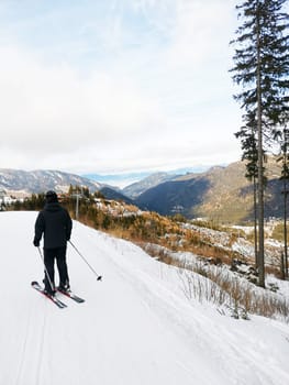 Beginner skier descends from a mountain on ski slope. View from the back of man in sports equipment. A vertical photo of man on slope, with mountains in the background