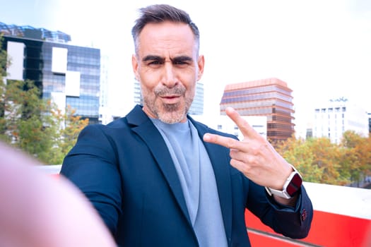 Stylish middle-aged man in a blazer taking a selfie while making a peace sign. He has a confident and playful expression. The background features a modern urban environment with buildings.