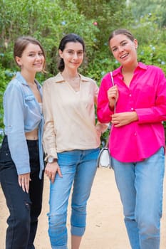 Three young women smiling and embracing each other while posing in a lush green park. They wear stylish casual outfits, expressing happiness, friendship, and joy in a natural outdoor setting.
