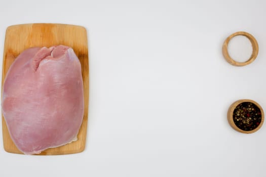 On the left is a piece of turkey fillet brisket, which lies on a wooden cutting board on a white background, on the right are seasonings in wooden bowls with space for text in the center. The concept of healthy eating, dietary and natural products, cooking. High quality photo