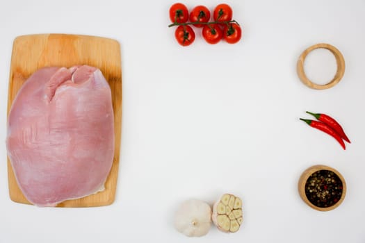 On the left is a piece of turkey fillet brisket lying on a wooden cutting board on a white background, with seasoning in wooden bowls on the edges, herbs and vegetables forming a place for the text in the center. The concept of healthy eating, dietary and natural products, cooking. High quality photo
