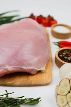 Vertical close-up photo of side view of a piece of turkey fillet brisket lying on a wooden cutting board on a white background surrounded by seasonings, herbs and vegetables. The concept of healthy eating, dietary and natural products, cooking. High quality photo