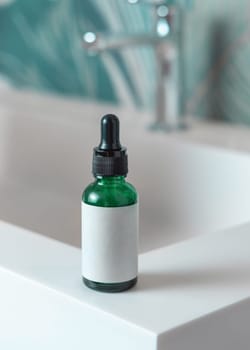 Green glass dropper bottle with pipette and blank label on washbasin with faucet in green tropical bathroom close up. Natural cosmetics - serum or essence. Daily beauty procedures