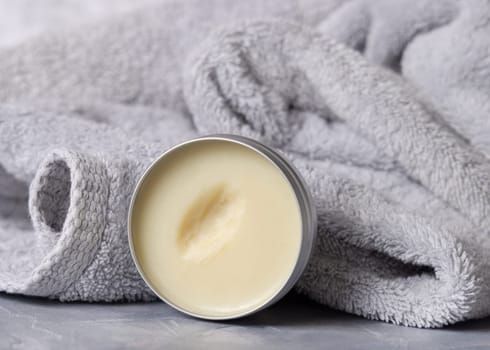 Opened body butter jar near light grey bath towel in bathroom, close up. Natural cosmetic product for body care, everyday beauty routine aand spa procedures